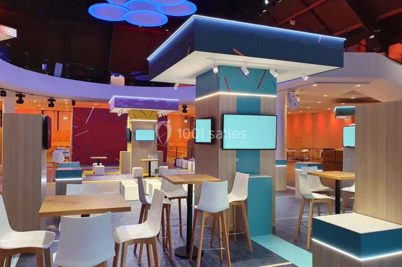 Espace intérieur moderne avec tables, chaises hautes et écrans, éclairé par des lumières colorées au plafond.