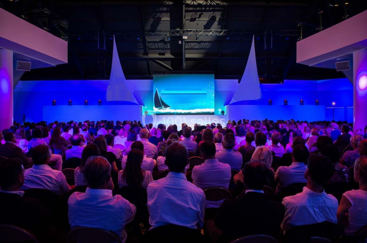 Un public assis dans une salle sombre regarde une projection lumineuse sur un écran, entouré d'éclairages bleus.