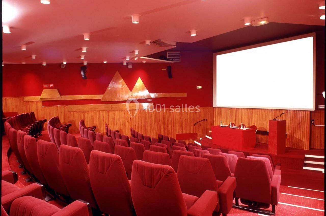 Salle de conférence avec sièges rouges, écran de projection blanc et éclairage au plafond.