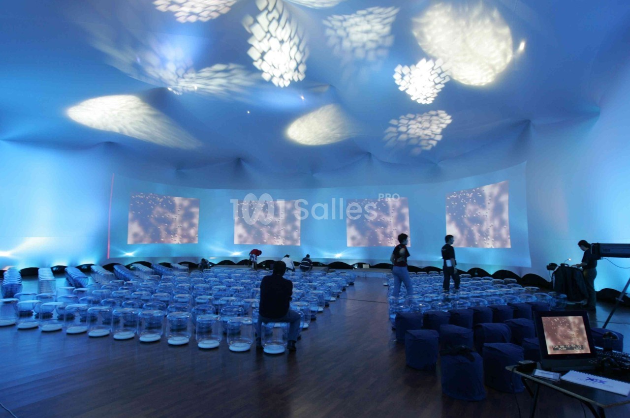 Salle immersive éclairée de projections lumineuses bleutées, avec des sièges transparents et quelques personnes présentes.
