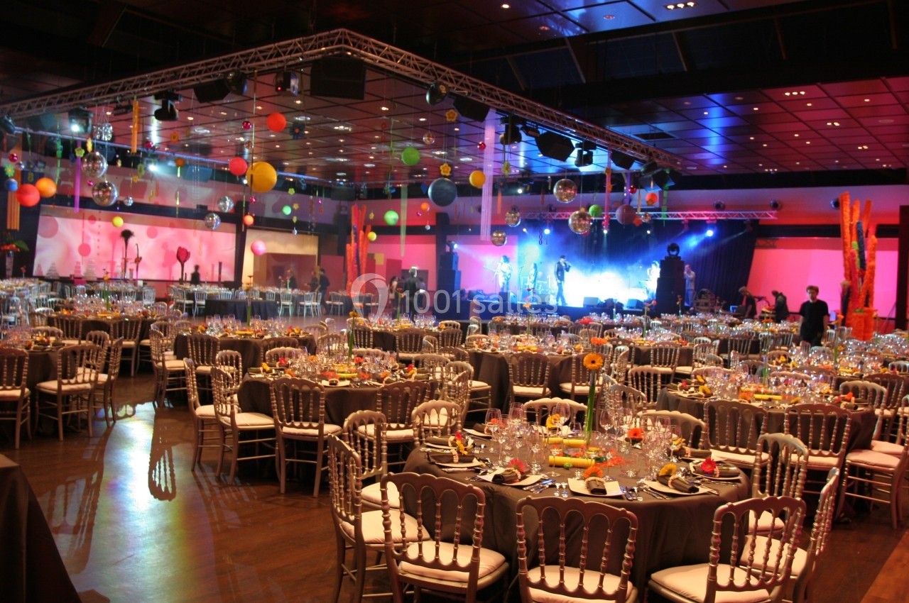 Salle de réception décorée avec des tables rondes dressées, éclairages colorés et scène en arrière-plan.