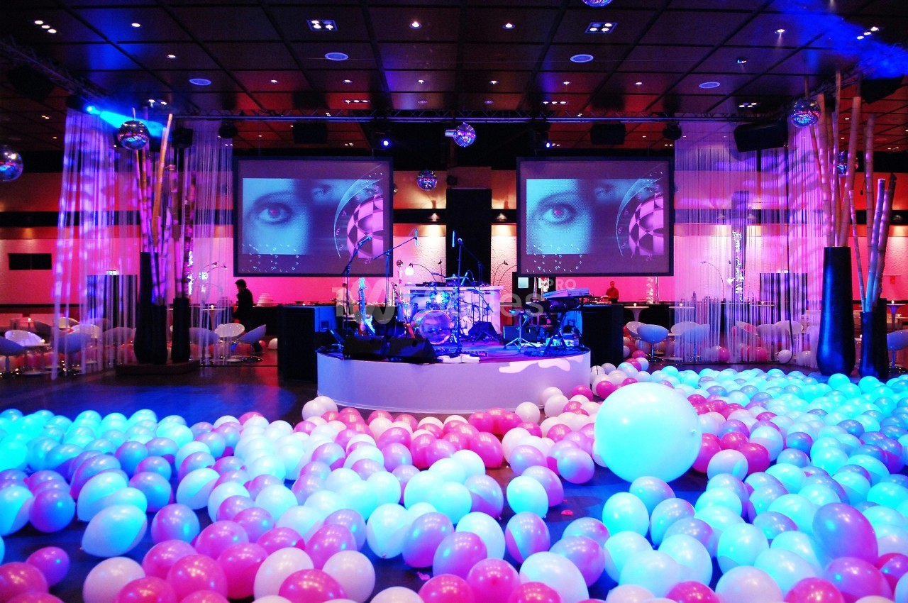 Scène de concert décorée avec des ballons roses et blancs, éclairage coloré et écrans projetant des images.