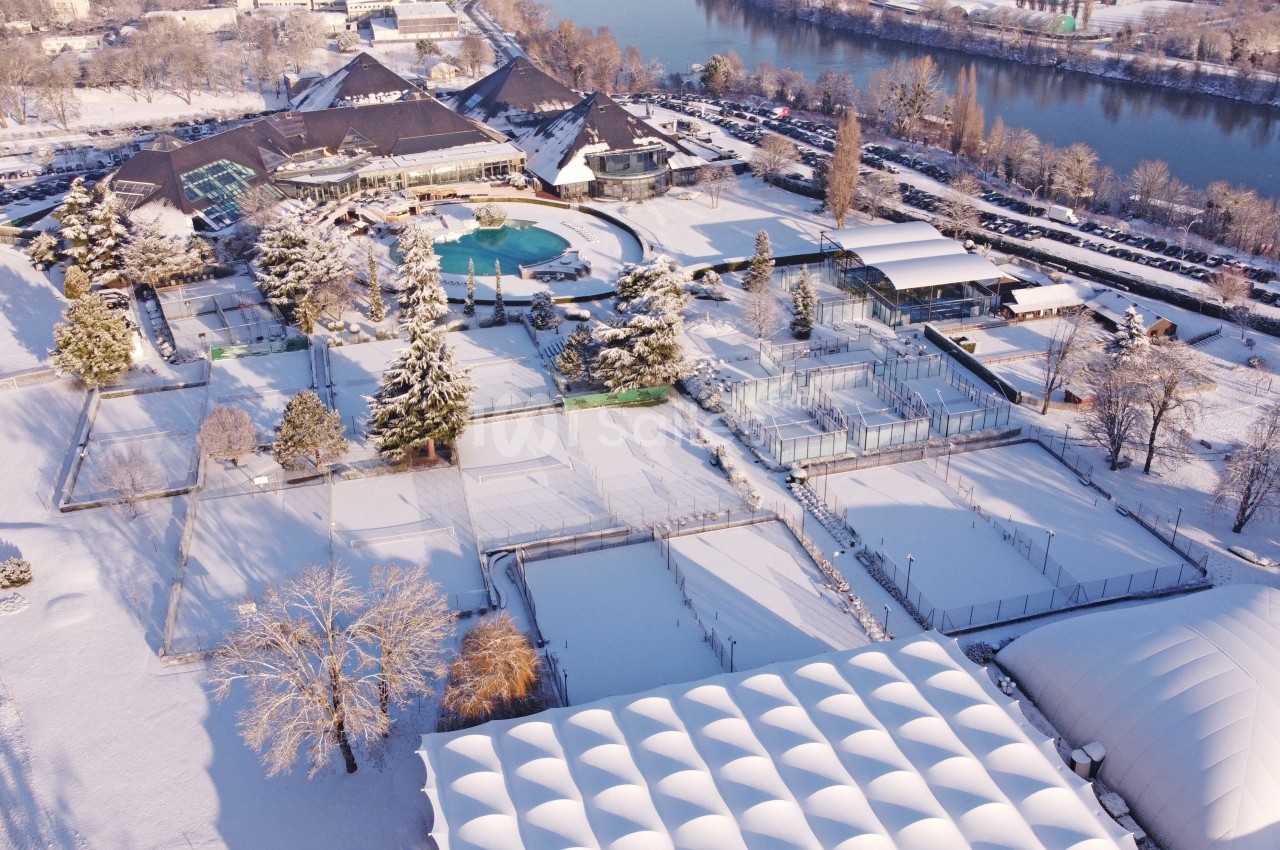 Vue aérienne d'un complexe sportif et de loisirs enneigé, avec piscine extérieure, terrains et bâtiments près d'une rivière.