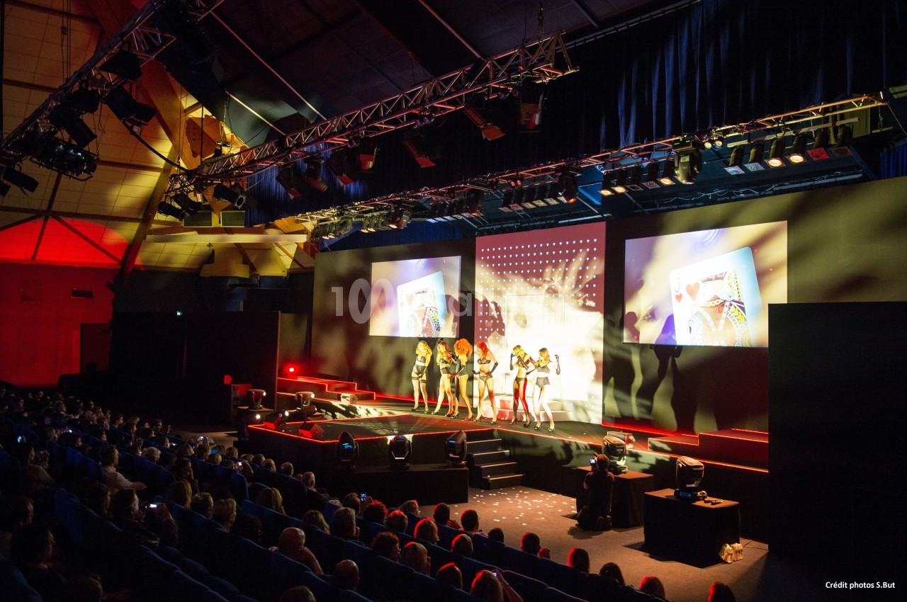 Spectacle sur scène avec des danseuses, projections visuelles et public assis dans une grande salle éclairée.