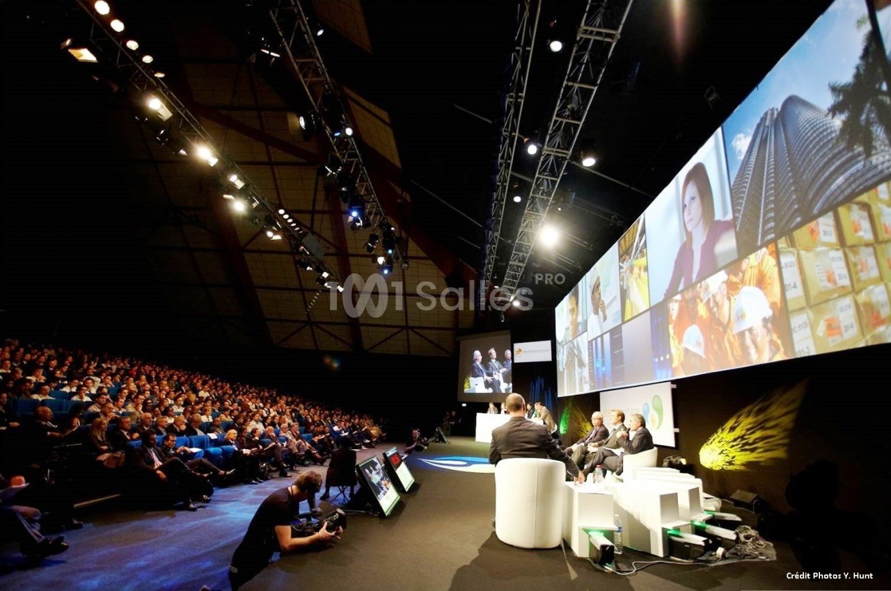 Vue d'une conférence avec un panel d'intervenants sur scène et un public assis dans une grande salle éclairée.
