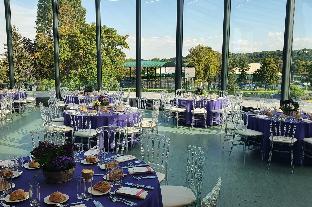 Salle de réception lumineuse avec tables rondes dressées, nappes violettes et chaises transparentes, vue sur un paysage…