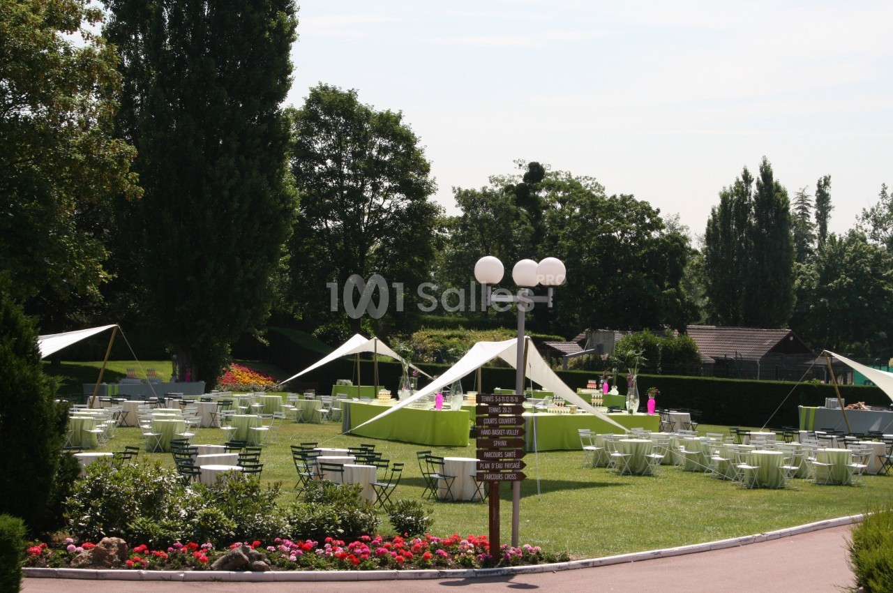 Tables et chaises disposées sur une pelouse avec des tonnelles blanches, entourées d'arbres et de parterres fleuris.
