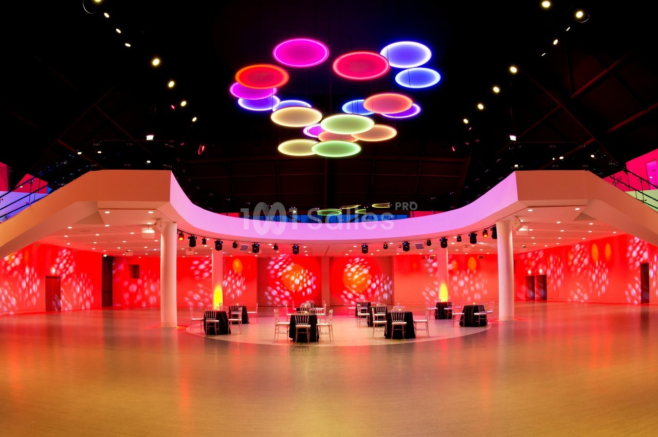 Salle spacieuse avec éclairage coloré au plafond, murs illuminés en rouge et tables disposées au centre.