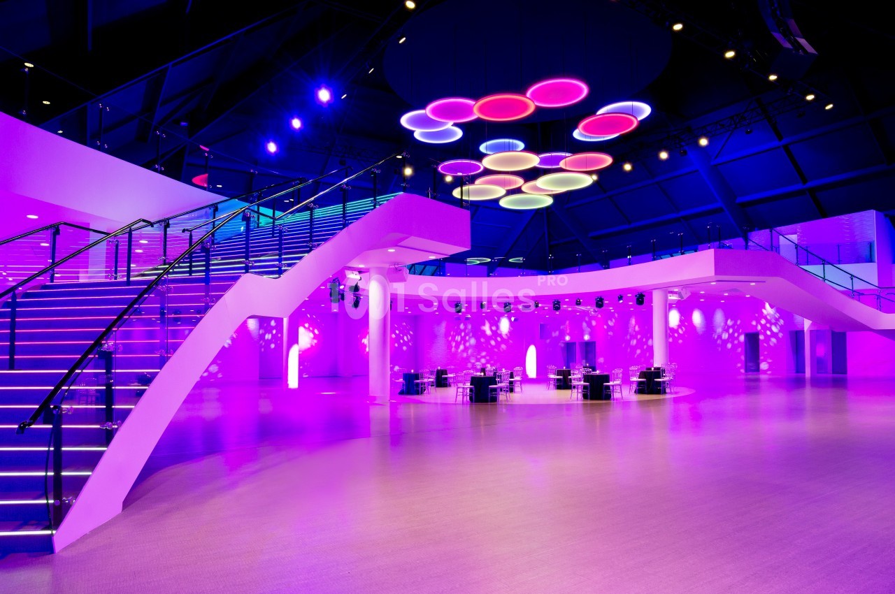 Salle moderne illuminée de lumières violettes avec escaliers, tables et plafonnier circulaire coloré.