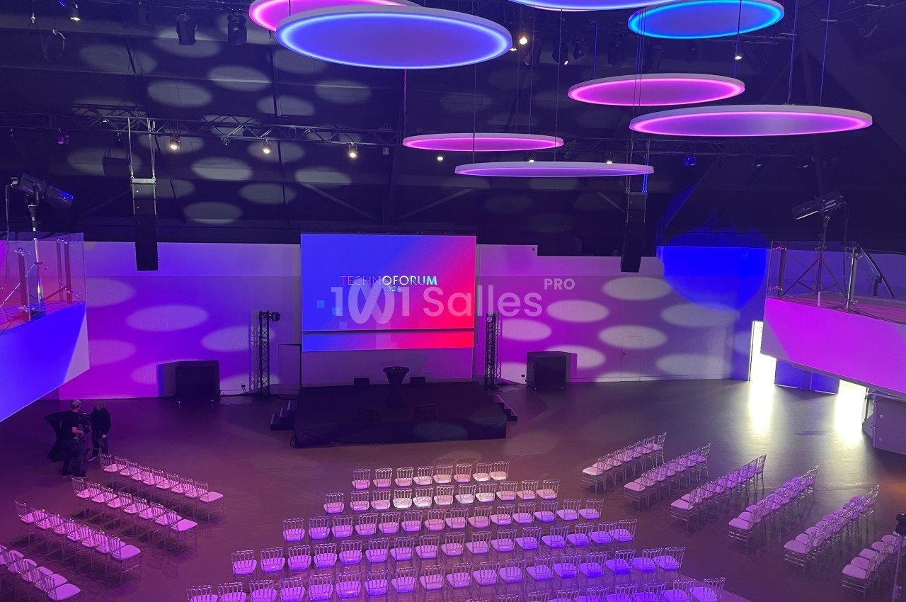 Salle de conférence moderne avec éclairage coloré, écran principal et rangées de chaises transparentes installées.