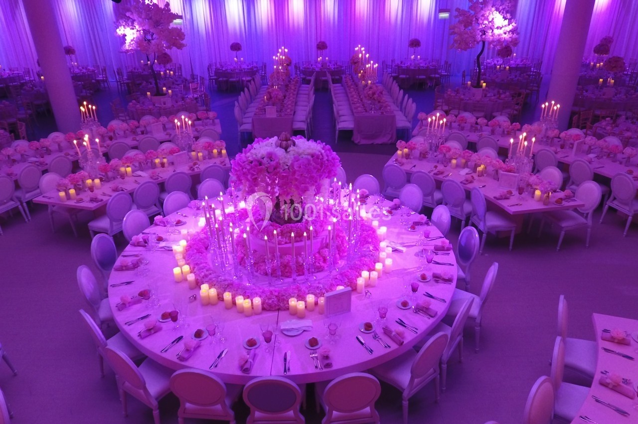 Salle de réception décorée en rose avec tables rondes, chandeliers et un grand centre de table floral illuminé.