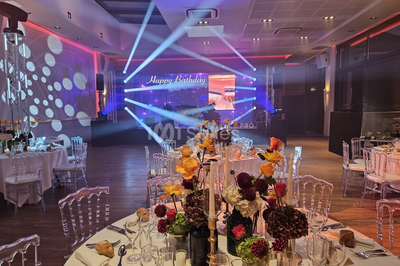 Salle de réception décorée pour un anniversaire, avec tables dressées, fleurs colorées et éclairage lumineux.