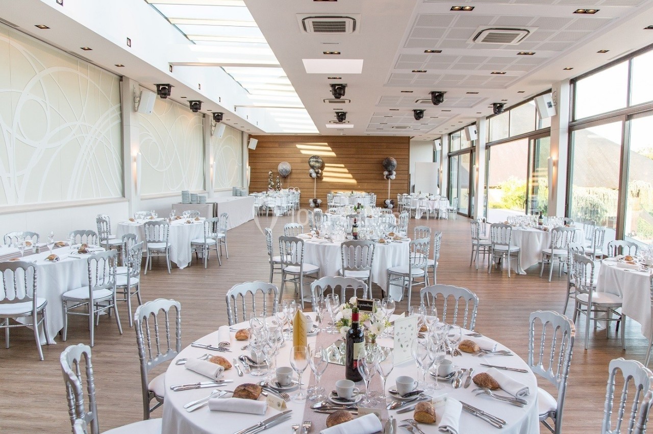 Salle de réception lumineuse avec tables rondes dressées, nappes blanches et chaises argentées.