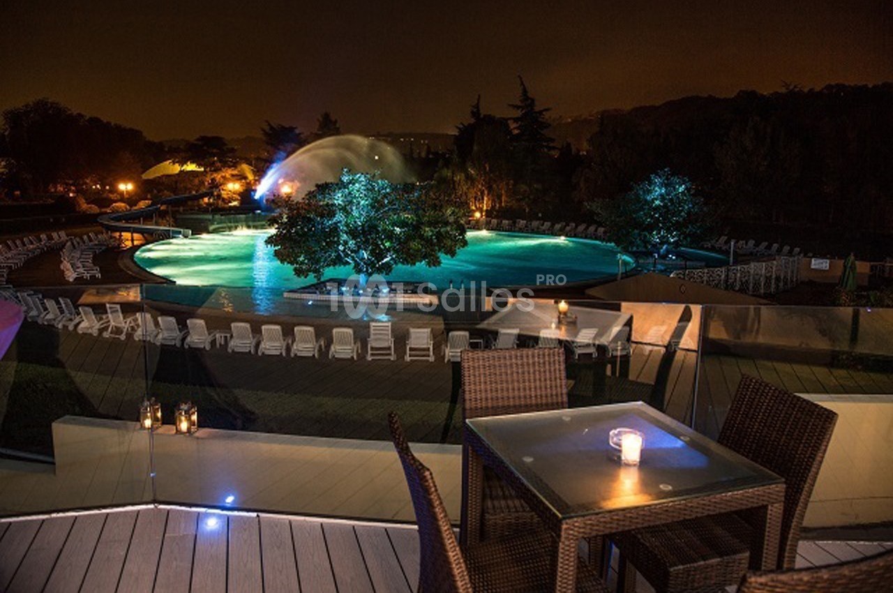 Piscine éclairée de nuit entourée de chaises longues, avec une table en premier plan et des arbres en arrière-plan.
