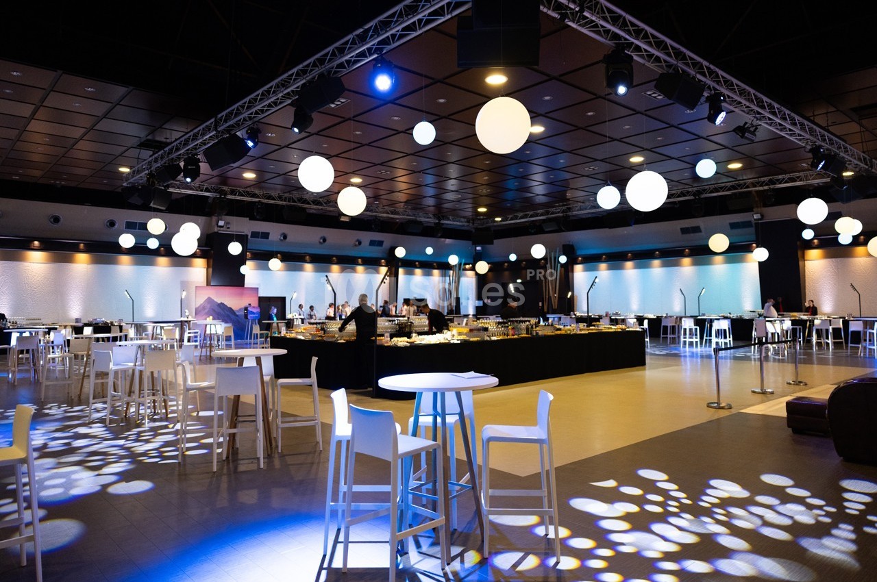 Salle de réception moderne avec tables hautes, éclairage décoratif et buffet au centre.