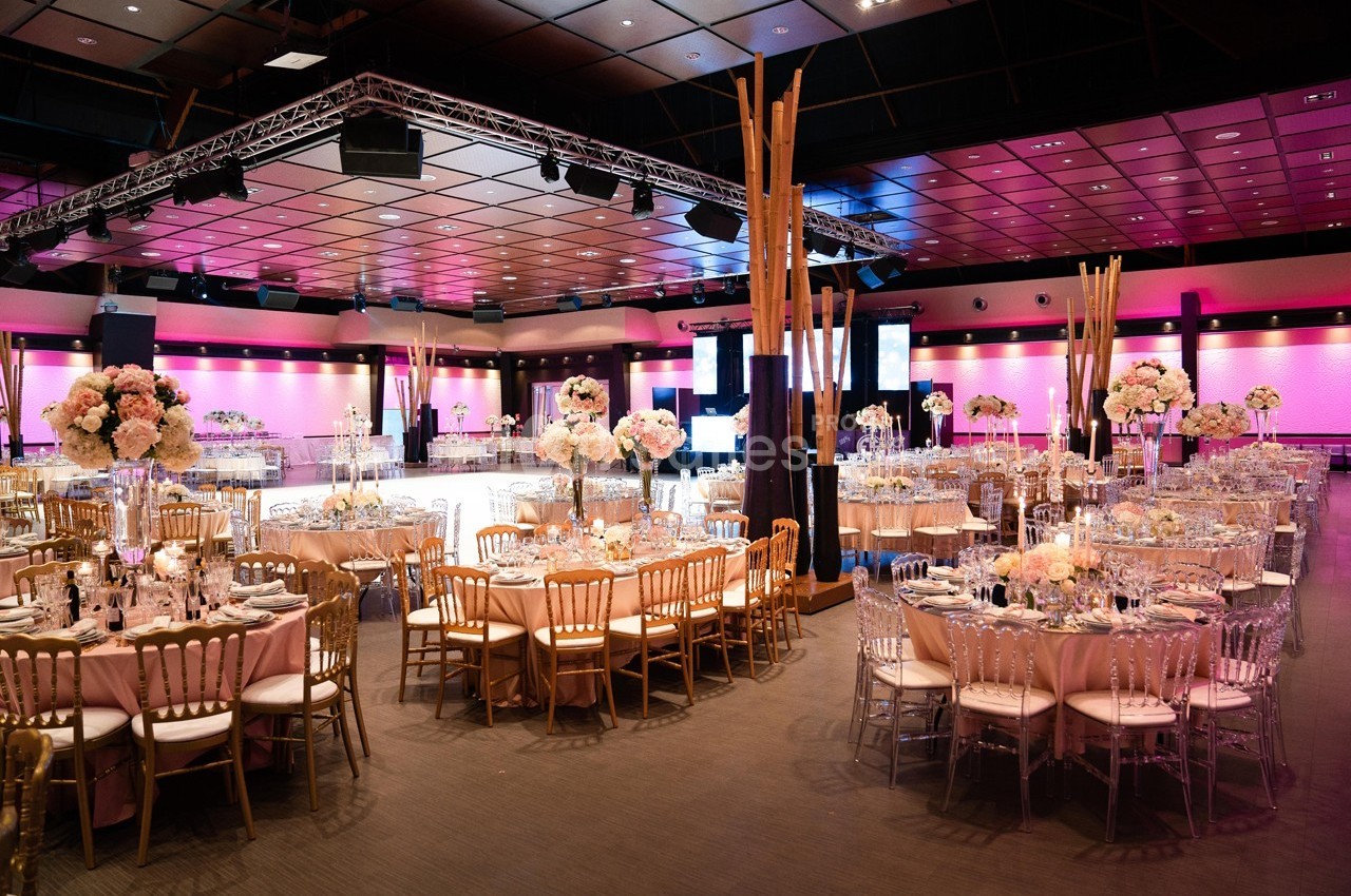 Salle de réception décorée avec des tables rondes, des chaises dorées et des centres de table fleuris sous un éclairage rose.