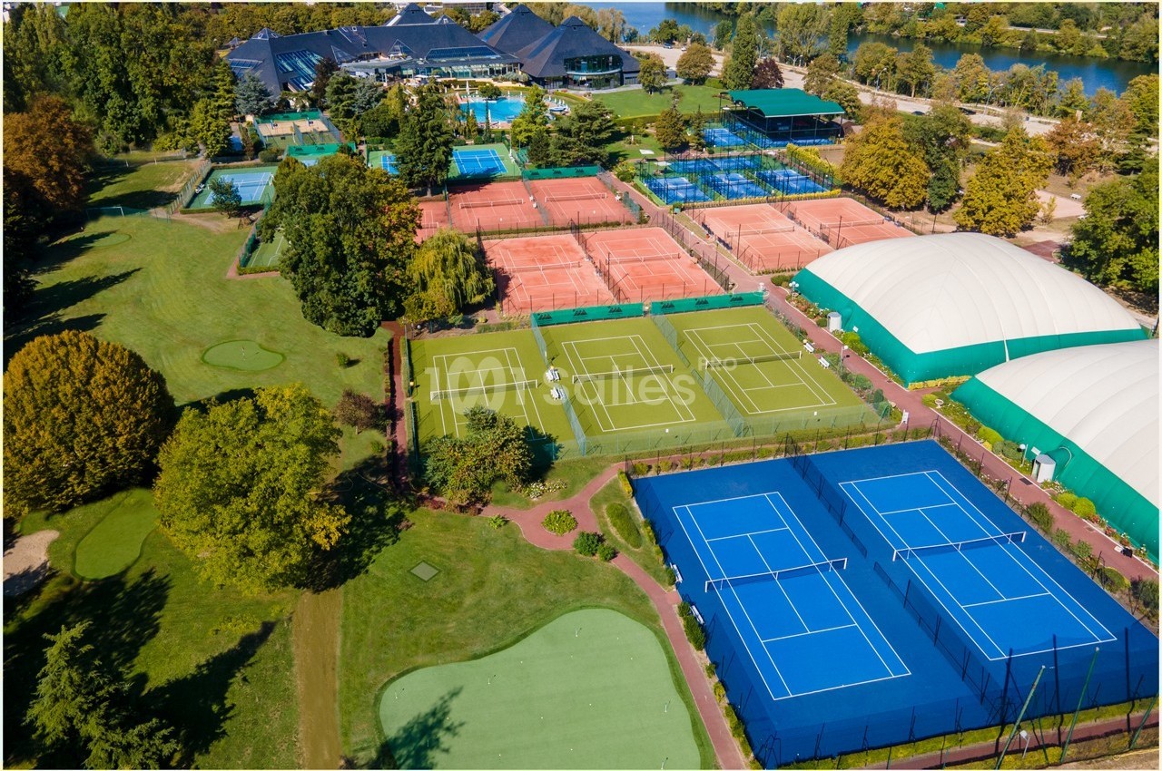 Vue aérienne d'un complexe sportif avec terrains de tennis en terre battue, gazon synthétique et courts couverts.
