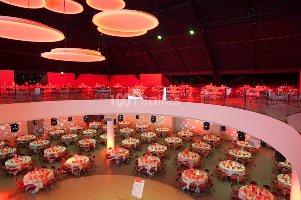 Salle de réception spacieuse avec des tables rondes dressées et une lumière rouge tamisée.