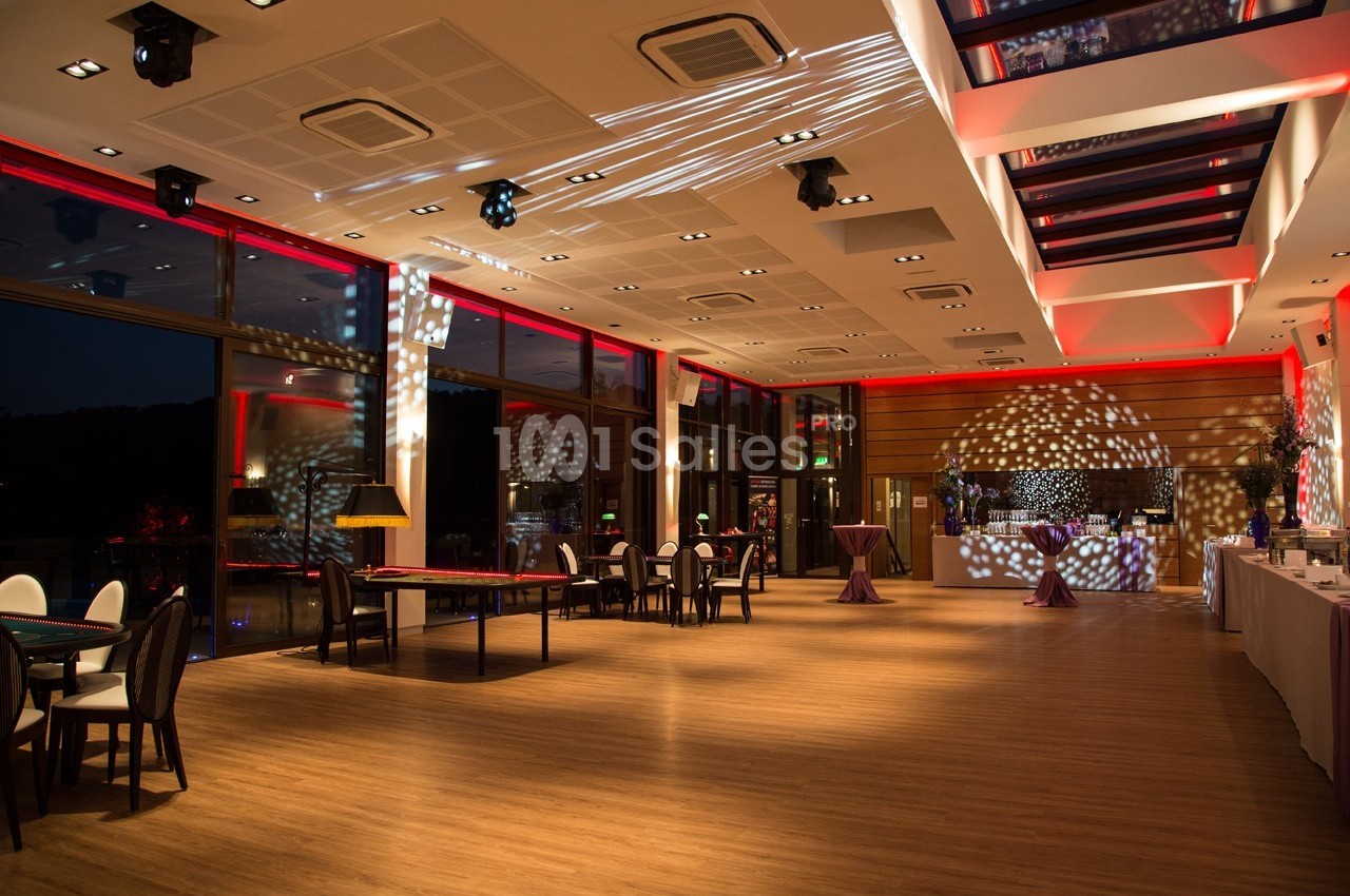 Salle de réception spacieuse avec éclairage tamisé, tables dressées et grandes baies vitrées donnant sur l'extérieur.