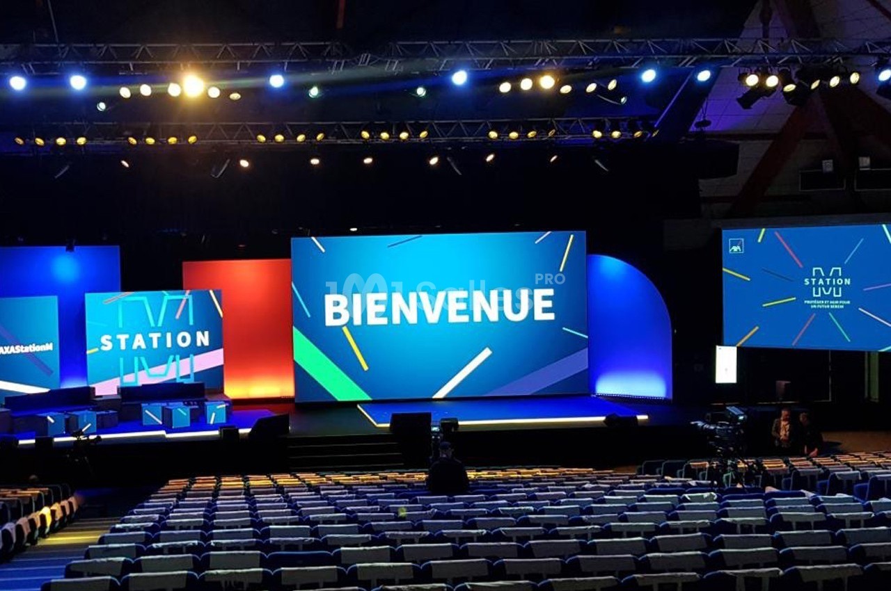 Scène d'un événement avec écrans affichant ’Bienvenue’ et ’Station’, entourée de sièges vides dans une grande salle.