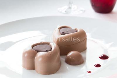 Dessert en forme de cœur au chocolat sur une assiette blanche, accompagné de coulis rouge et d'une verrine en arrière-plan.