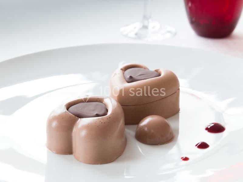 Dessert en forme de cœur au chocolat sur une assiette blanche, accompagné de coulis rouge et d'une verrine en arrière-plan.