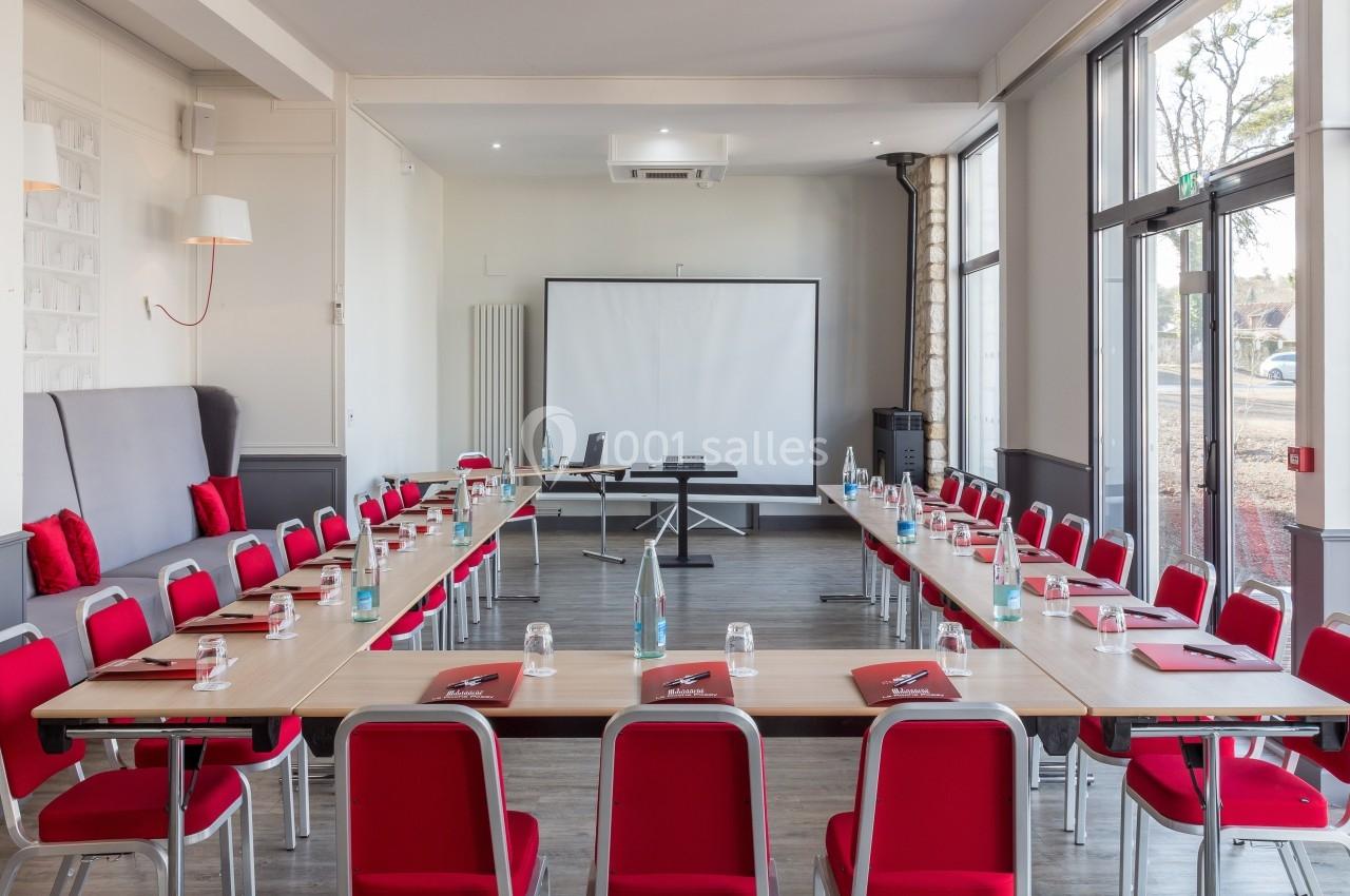 Salle de réunion lumineuse avec tables disposées en U, chaises rouges, écran de projection et grandes fenêtres.