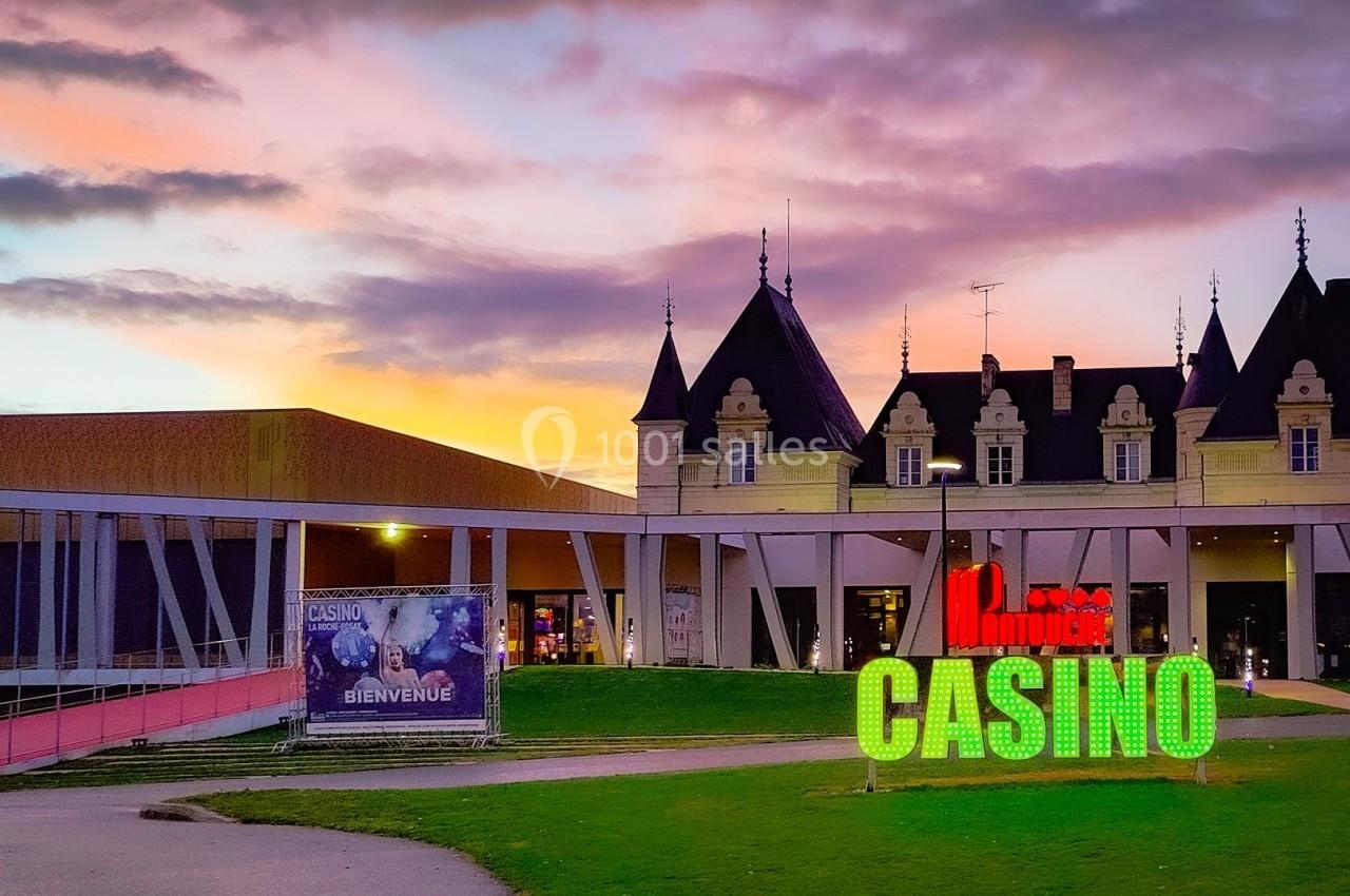 Location salle La Roche-Posay (Vienne) - Casino de la Roche Posay #2 Façade d'un casino avec enseigne lumineuse, devant un bâtiment de style château au coucher du soleil.