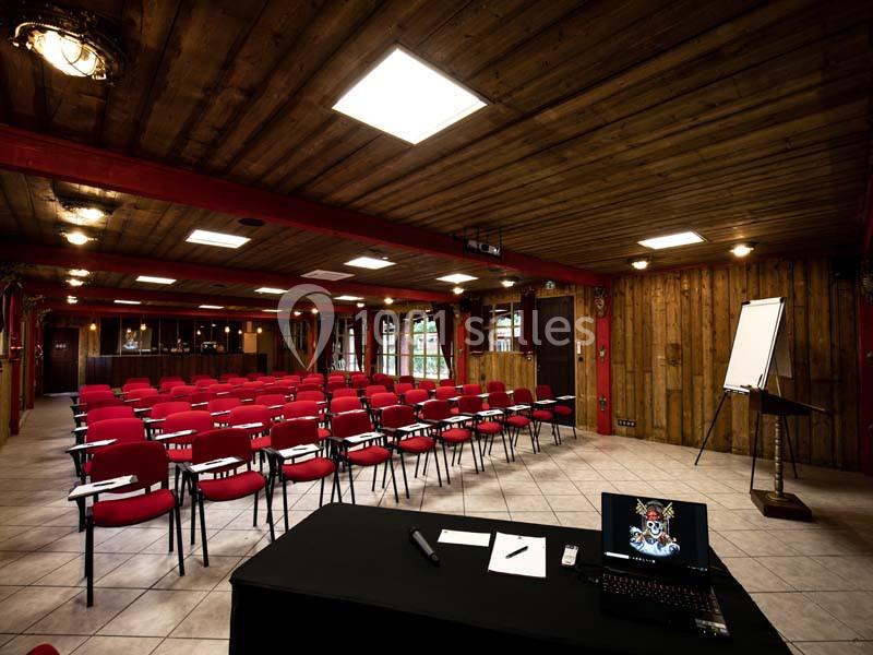 Salle de conférence avec rangées de chaises rouges, un bureau équipé d'un ordinateur portable et un tableau blanc.