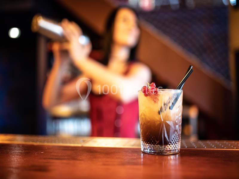 Un cocktail décoré de groseilles et d'une paille noire posé sur un bar, avec une personne préparant un mélange en arrière…