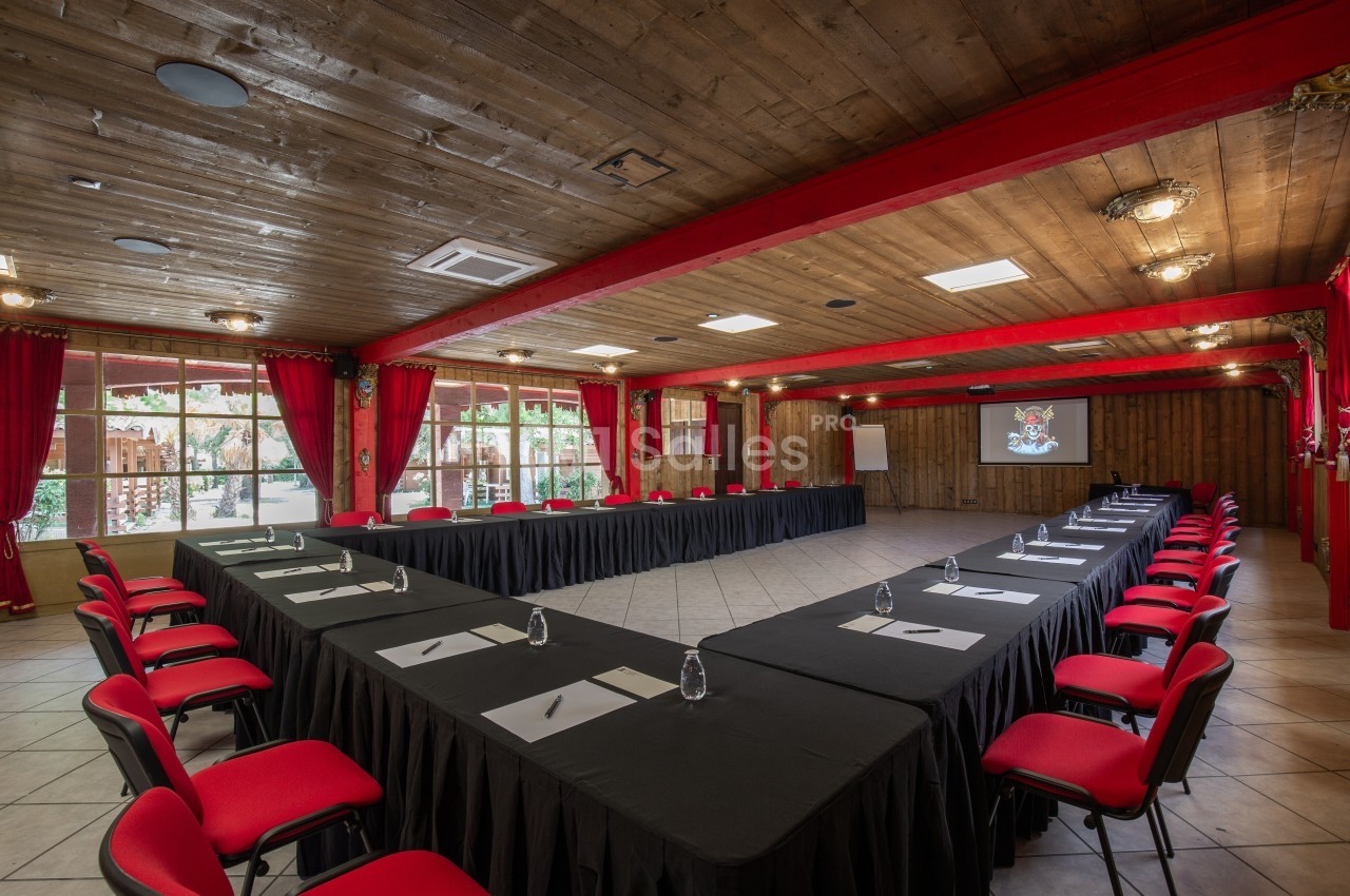 Salle de réunion spacieuse avec tables disposées en U, chaises rouges, grandes fenêtres et écran de projection.