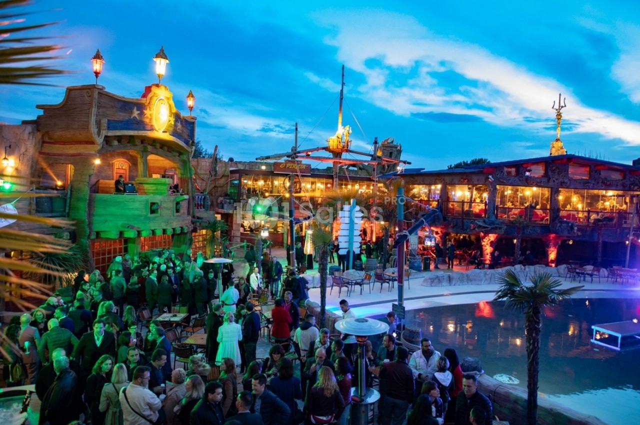 Foule rassemblée dans un lieu extérieur animé, décoré sur le thème des pirates, avec éclairages colorés et piscine.