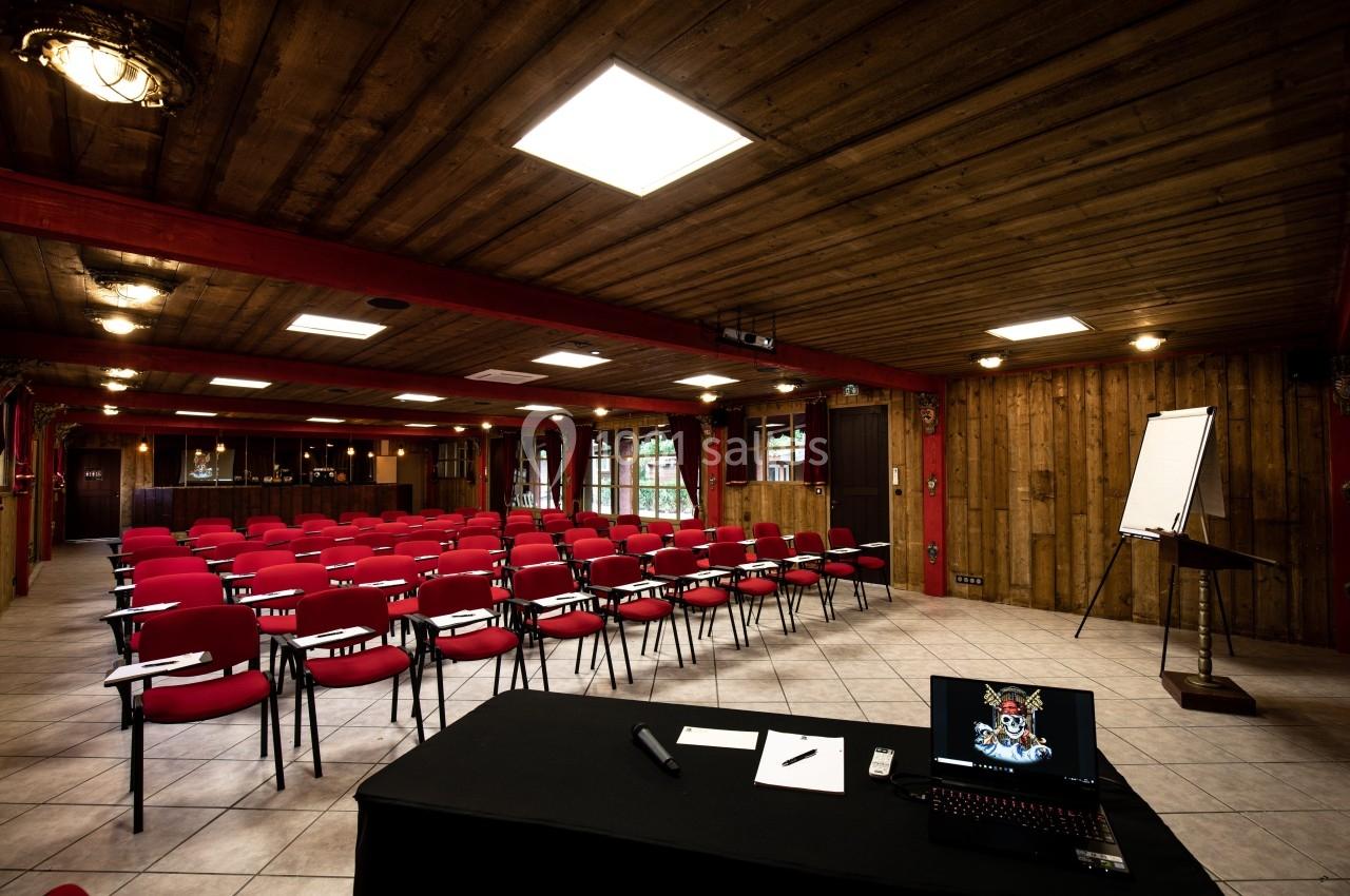 Salle de conférence avec des chaises rouges alignées, un bureau avec ordinateur portable et un tableau de présentation.