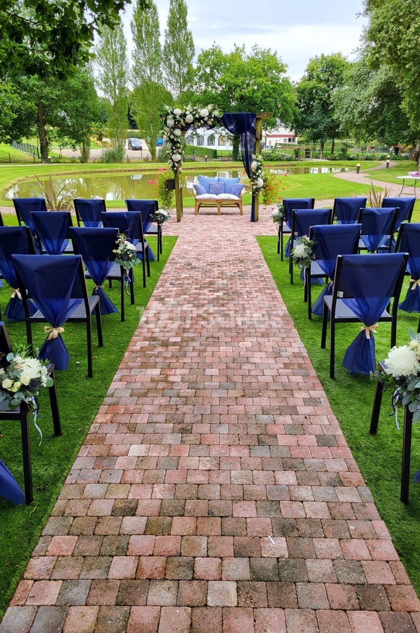 Allée en briques menant à une arche décorée de fleurs blanches et bleues, entourée de chaises ornées de rubans.
