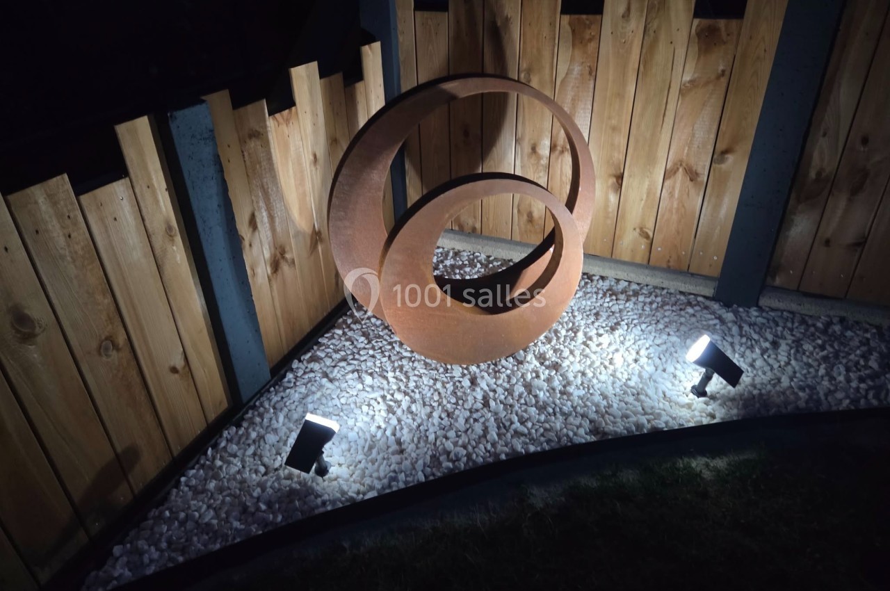 Sculpture métallique circulaire éclairée dans un jardin, entourée de graviers blancs et d'une clôture en bois.