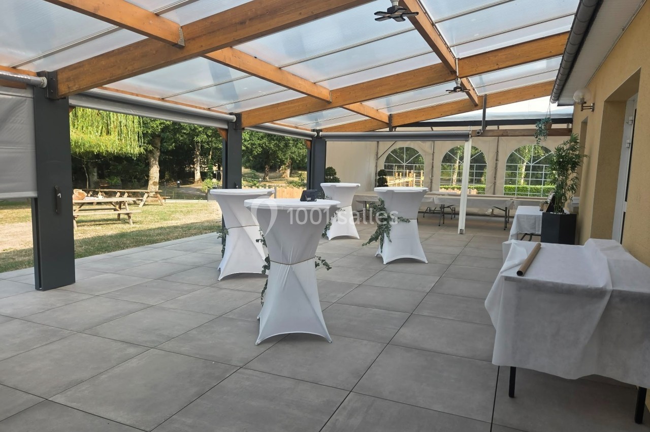 Terrasse couverte avec tables hautes drapées de blanc, entourée de verdure et éclairée par une lumière naturelle.