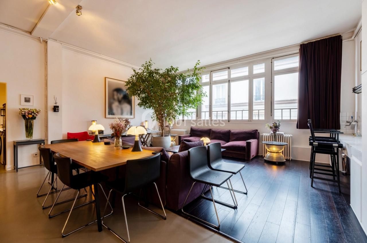 Salle de séjour lumineuse avec table à manger en bois, chaises noires, canapé violet et grande plante près des fenêtres.