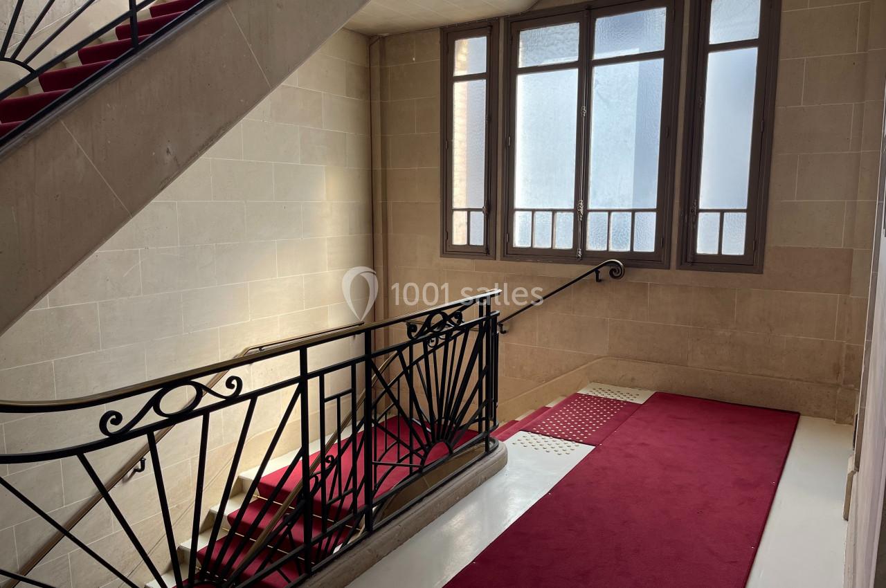 Escalier intérieur avec rampe en fer forgé, tapis rouge et fenêtre laissant entrer la lumière naturelle.