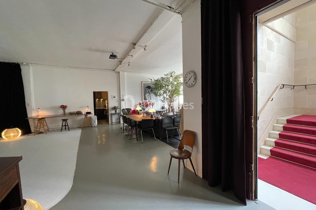 Espace intérieur lumineux avec mobilier éclectique, plantes, horloge murale et escalier à tapis rouge visible à droite.