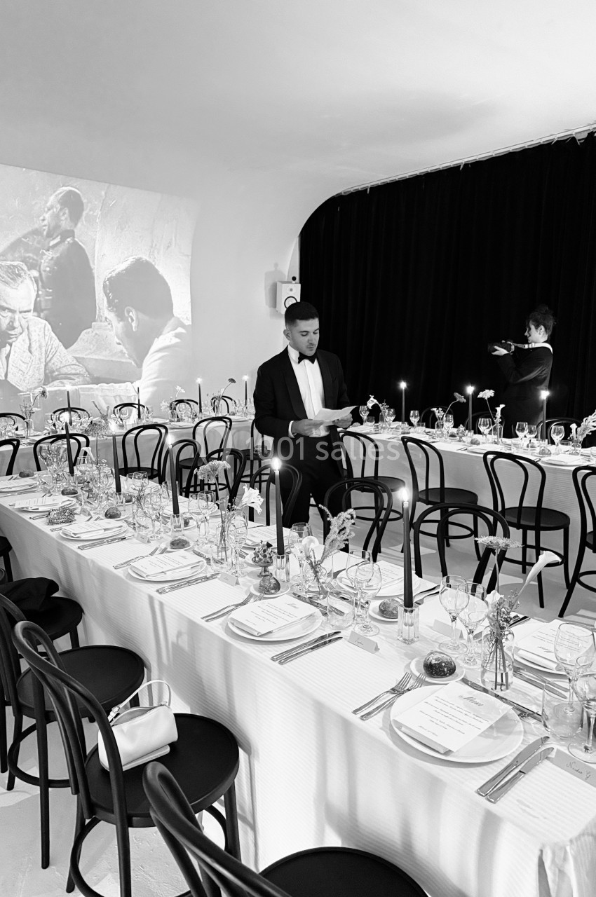 Salle de réception élégante avec tables dressées, chandeliers allumés et un serveur en costume ajustant les détails.