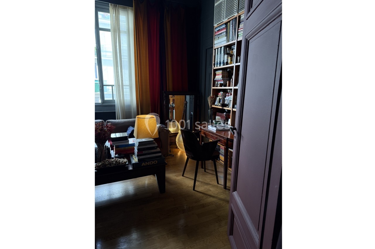 Location salle Paris 20 (Paris) - Studio 7 #29 Pièce sombre avec bibliothèque, bureau en bois, lampe allumée et fauteuil, près d'une fenêtre avec rideaux.