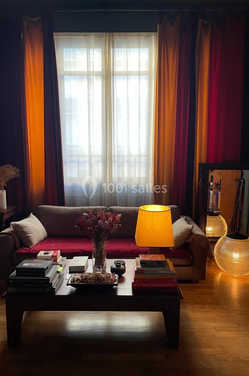 Location salle Paris 20 (Paris) - Studio 7 #5 Salon avec canapé beige, table basse chargée de livres, lampe allumée et rideaux colorés devant une fenêtre.