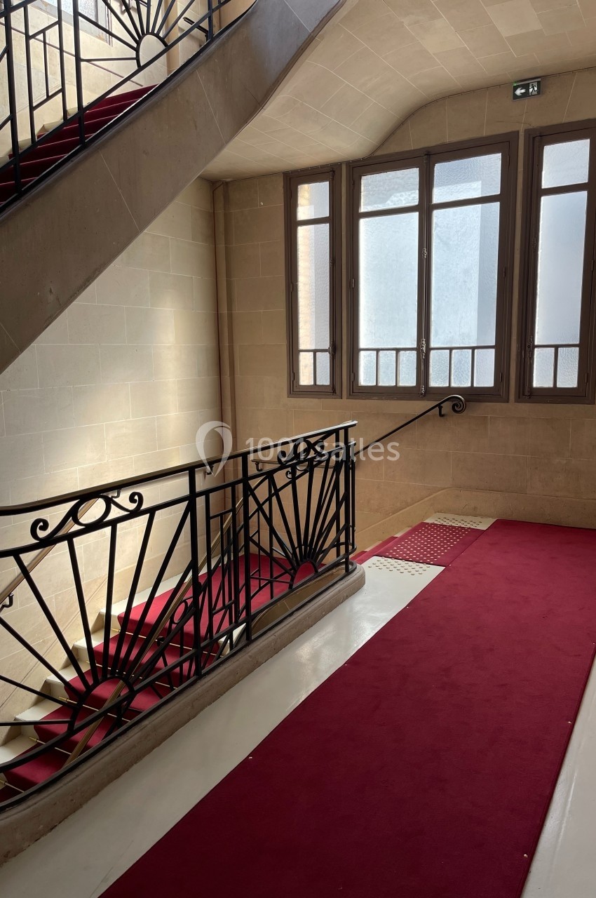 Escalier intérieur en pierre avec rampe en fer forgé, tapis rouge et grande fenêtre laissant entrer la lumière naturelle.