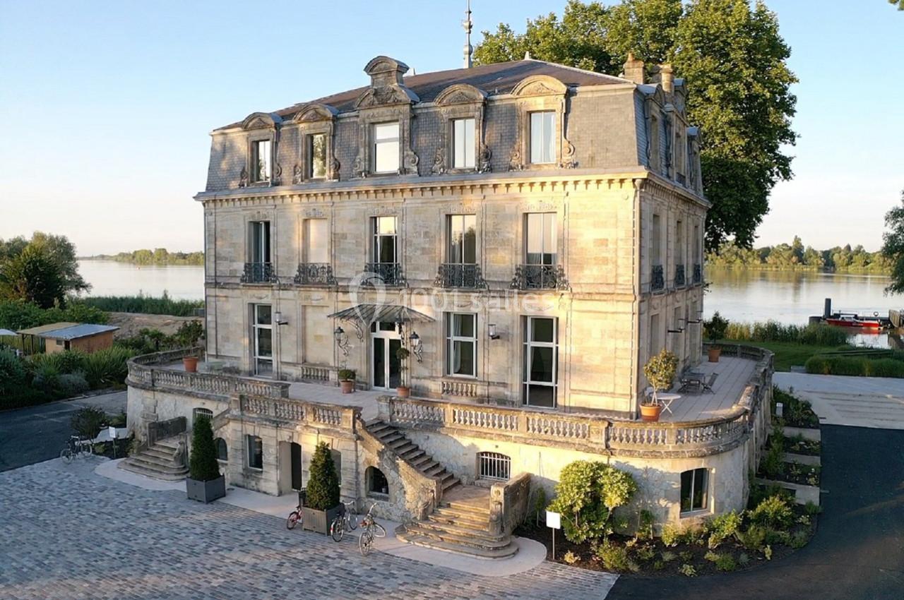Façade d'un bâtiment ancien en pierre avec balcons, entouré de végétation et situé près d'un cours d'eau.