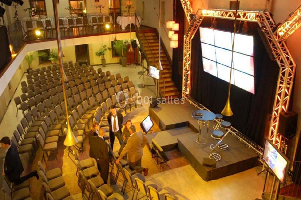 Salle de conférence avec sièges alignés, scène équipée d'écrans et de projecteurs, quelques personnes discutant.