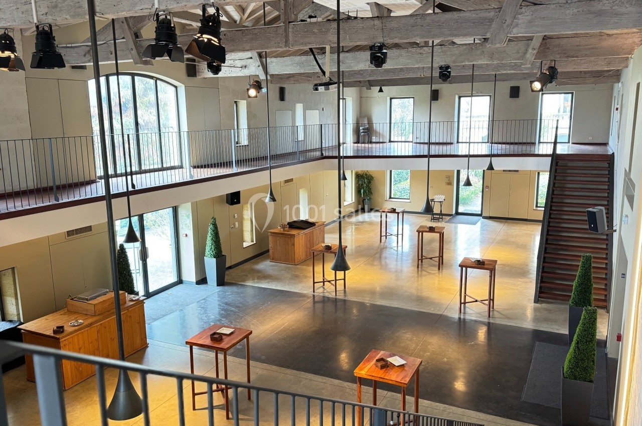 Salle spacieuse et lumineuse avec mezzanine, tables hautes en bois, grandes fenêtres et poutres apparentes.