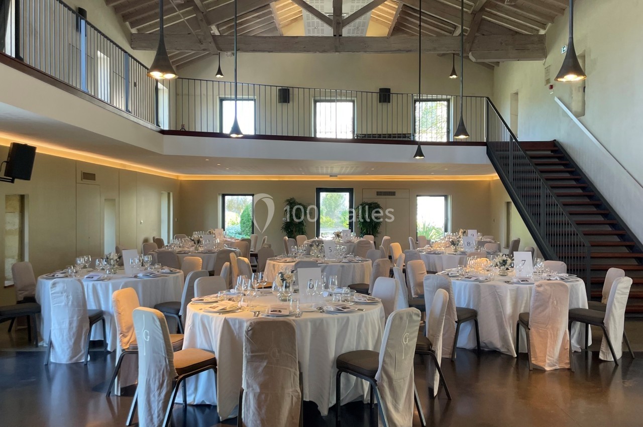 Salle de réception lumineuse avec tables rondes dressées, chaises couvertes et mezzanine en bois visible en arrière-plan.