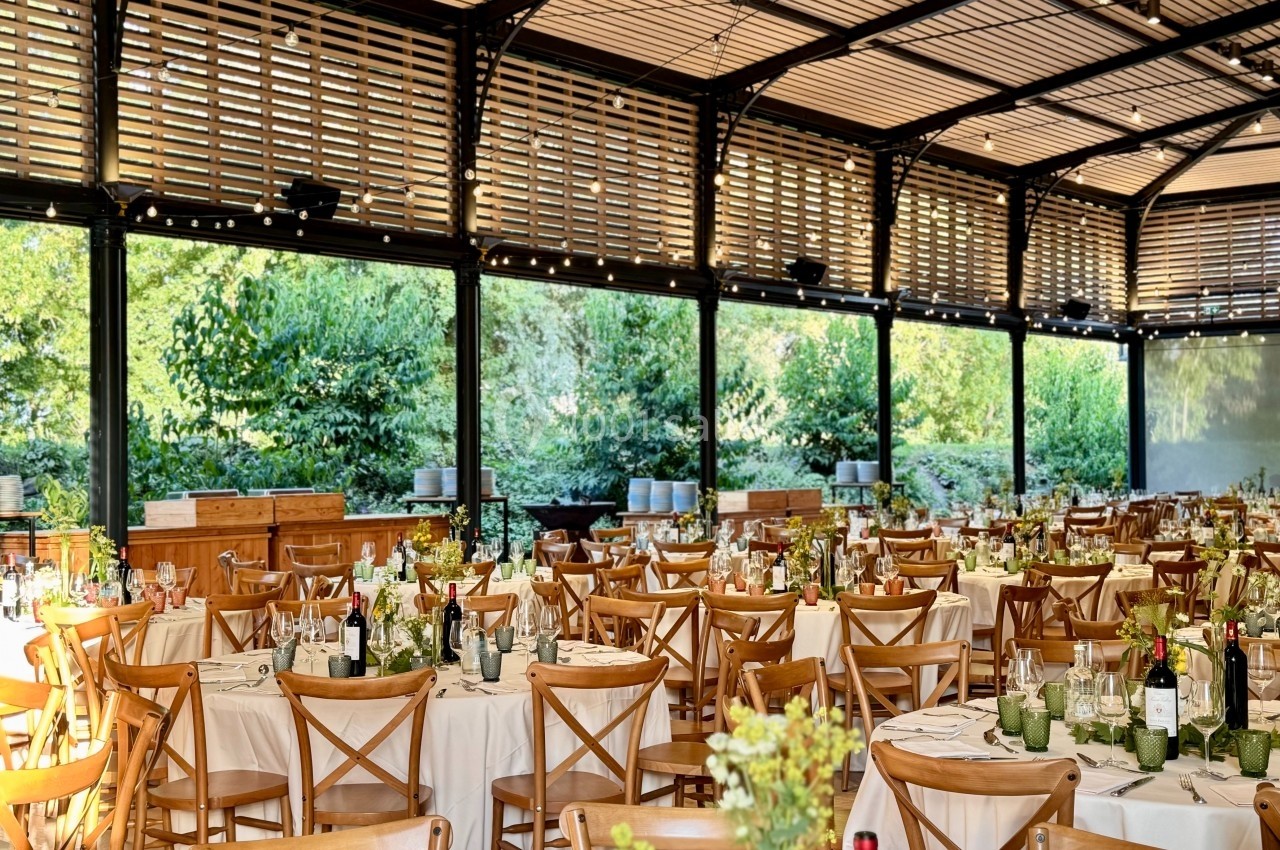 Salle de réception lumineuse avec tables dressées, chaises en bois et vue sur un jardin verdoyant à travers de grandes baies…