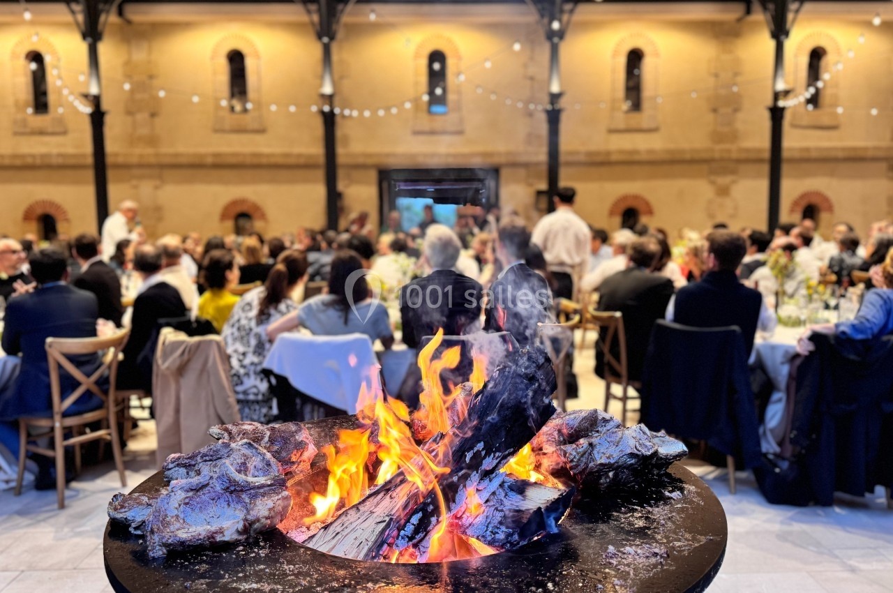 Brasero allumé au premier plan, avec des convives assis à des tables dans une cour éclairée en arrière-plan.