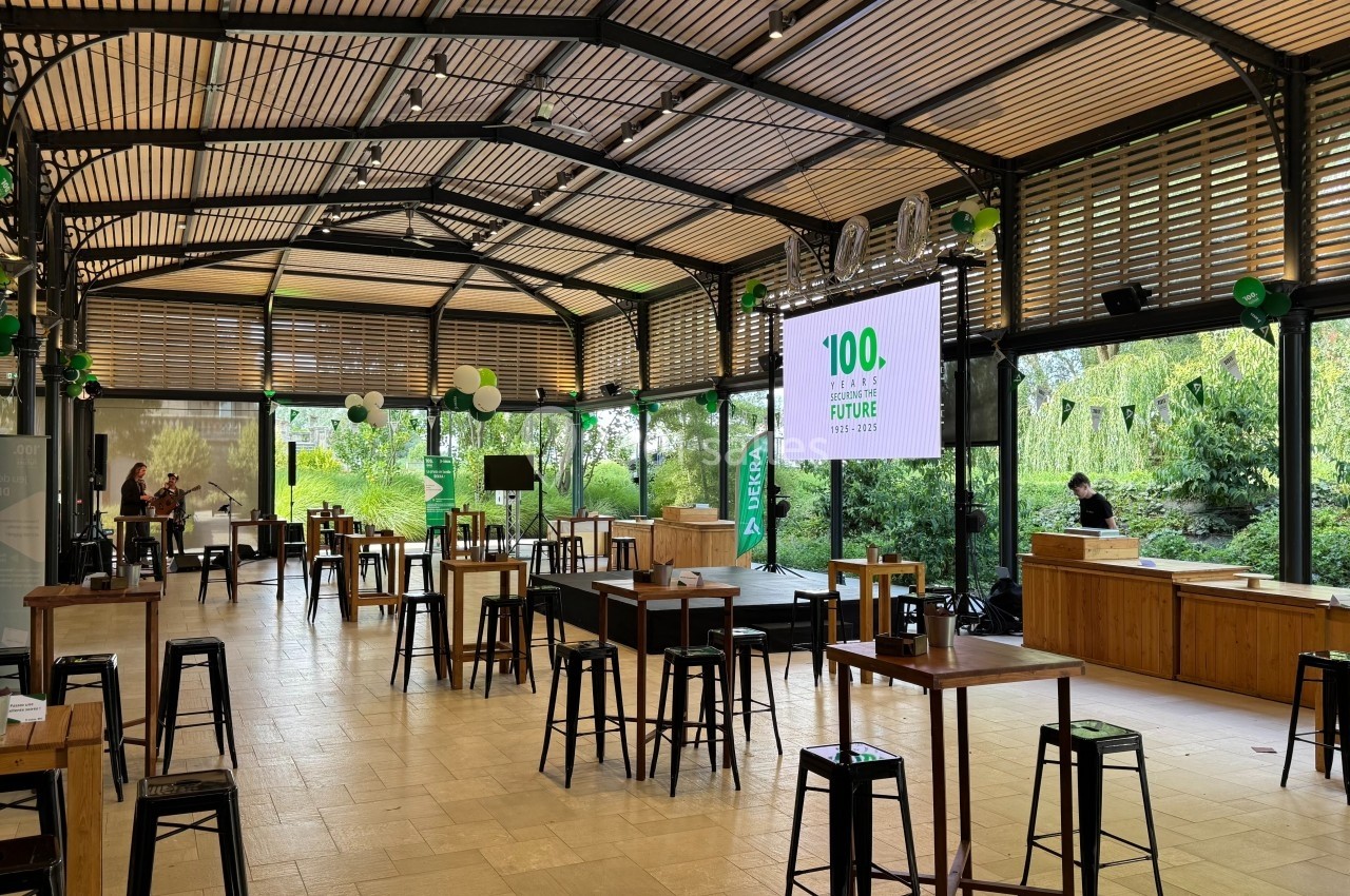 Salle lumineuse avec tables hautes, tabourets noirs et décorations vertes, préparée pour un événement ou une réception.